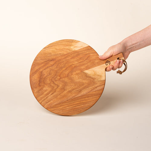 A hand holds a small round cherry serving board with a short handle and leather hanging loop, shown held against a warm cream background.