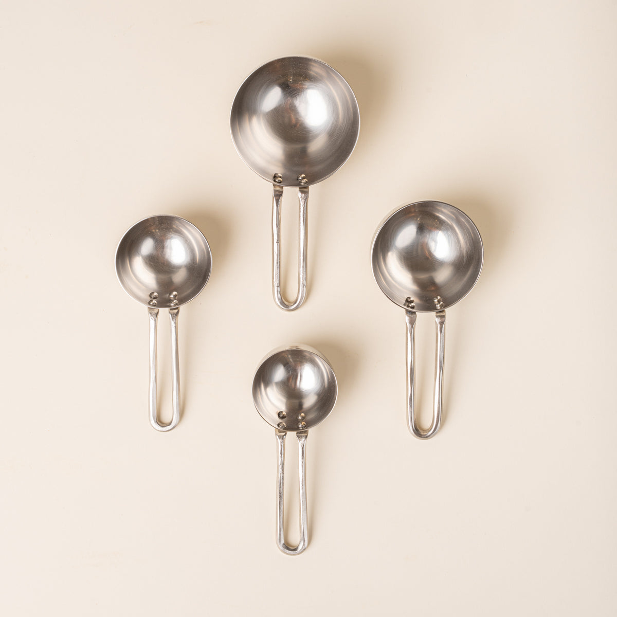Four forged measuring cups in graduated sizes, arranged in a loose diamond formation on a warm cream background.