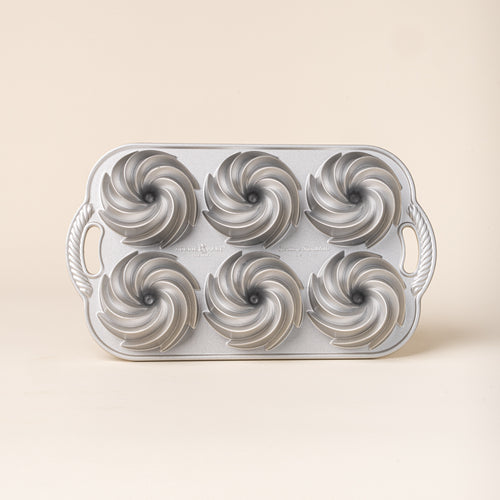 Silver metal baking pan with six spiral-shaped mini Bundt cake molds and decorative handles on each side.