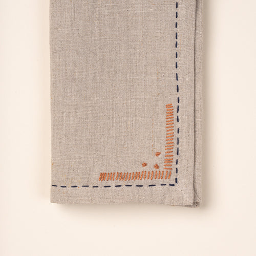 Close up of a corner of a folded natural linen napkins featuring dark blue running-stitch borders and small rust-orange embroidered corner details.