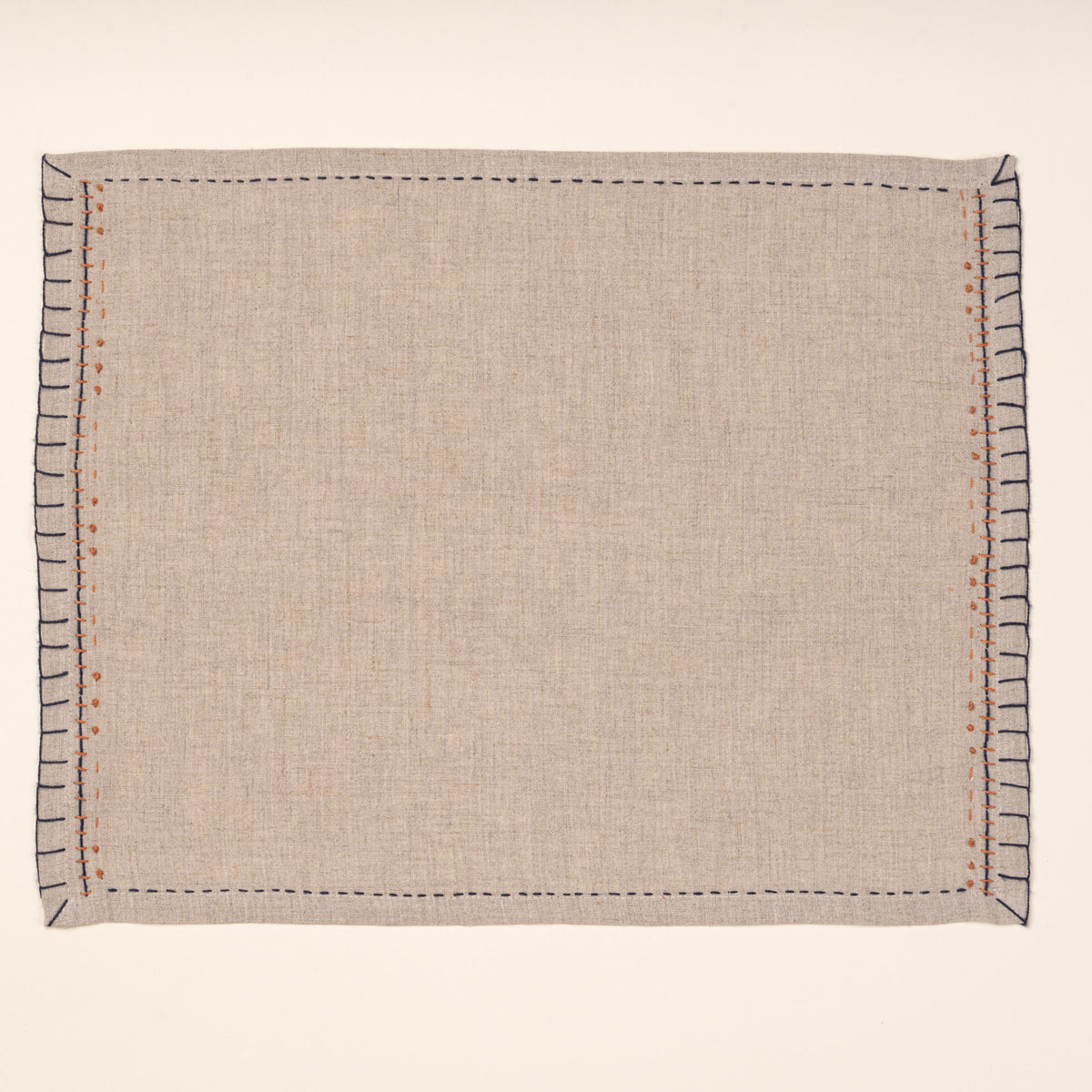 A natural flax-colored linen placemat with hand-stitched navy borders and rust-orange accent embroidery, photographed against a warm off-white background.