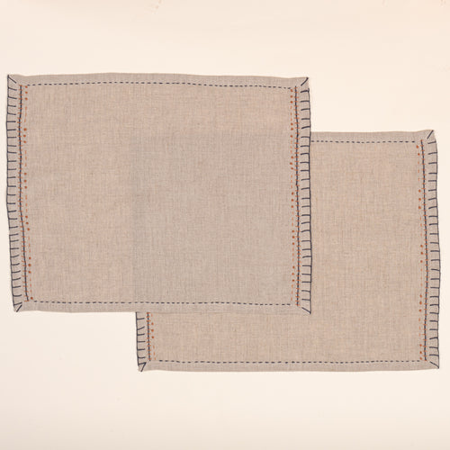 Two overlapping natural flax-colored linen placemats with hand-stitched navy borders and rust-orange accent embroidery, photographed against a warm off-white background.