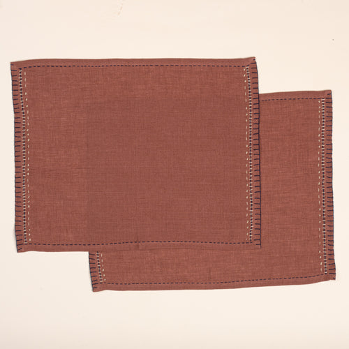 Two overlapping rust-brown linen placemats with hand-stitched dark blue borders and contrasting cream decorative embroidery, photographed against a warm off-white background.