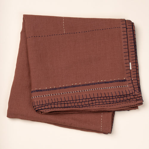 Folded rust-brown linen tablecloth with hand-stitched navy and cream detailing and a decorative geometric border.
