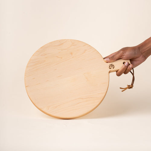 A hand holds a small round maple serving board with a short handle and leather hanging loop, shown held against a warm cream background.