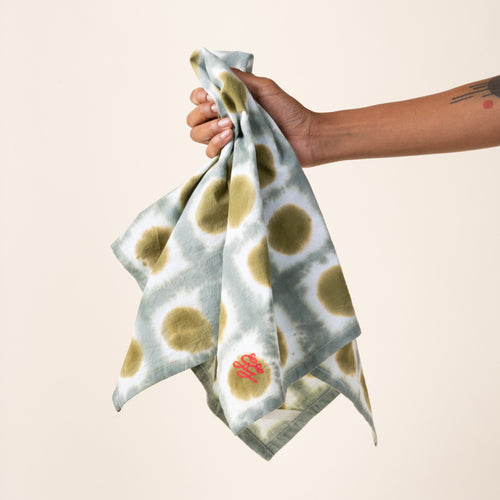 A hand holding out a tie-dye tea towel in a green colorway with a red embroidered corner
