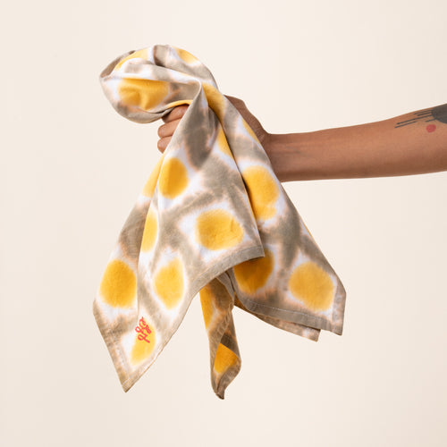 A hand holding out a tie-dye tea towel in a yellow colorway with a red embroidered corner