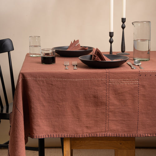 Rust-brown linen tablecloth with hand-stitched details, styled with dark dinnerware, matching napkins, glassware, and candles.