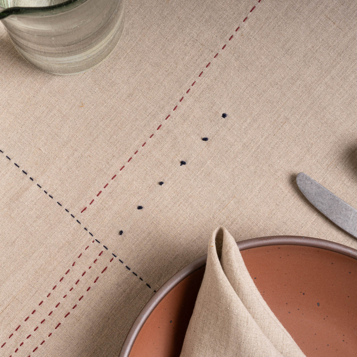 Closeup of natural linen tablecloth with hand-stitched navy and rust details, styled with warm ceramics and glassware.