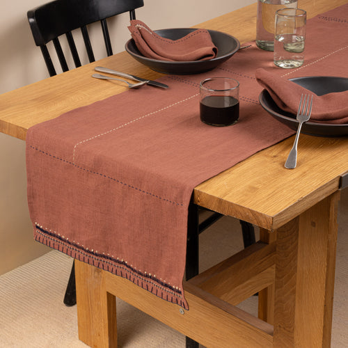 A rust-brown linen table runner styled across a warm oak dining table with matching napkins, dark ceramic dinnerware, simple glassware, and silver flatware.