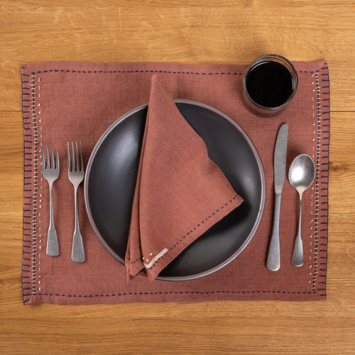 A rust-brown linen placemat and coordinating napkin with dark blue running-stitch borders and white embroidered details, styled with a dark ceramic plate, silver flatware, and a glass on a warm wood tabletop.