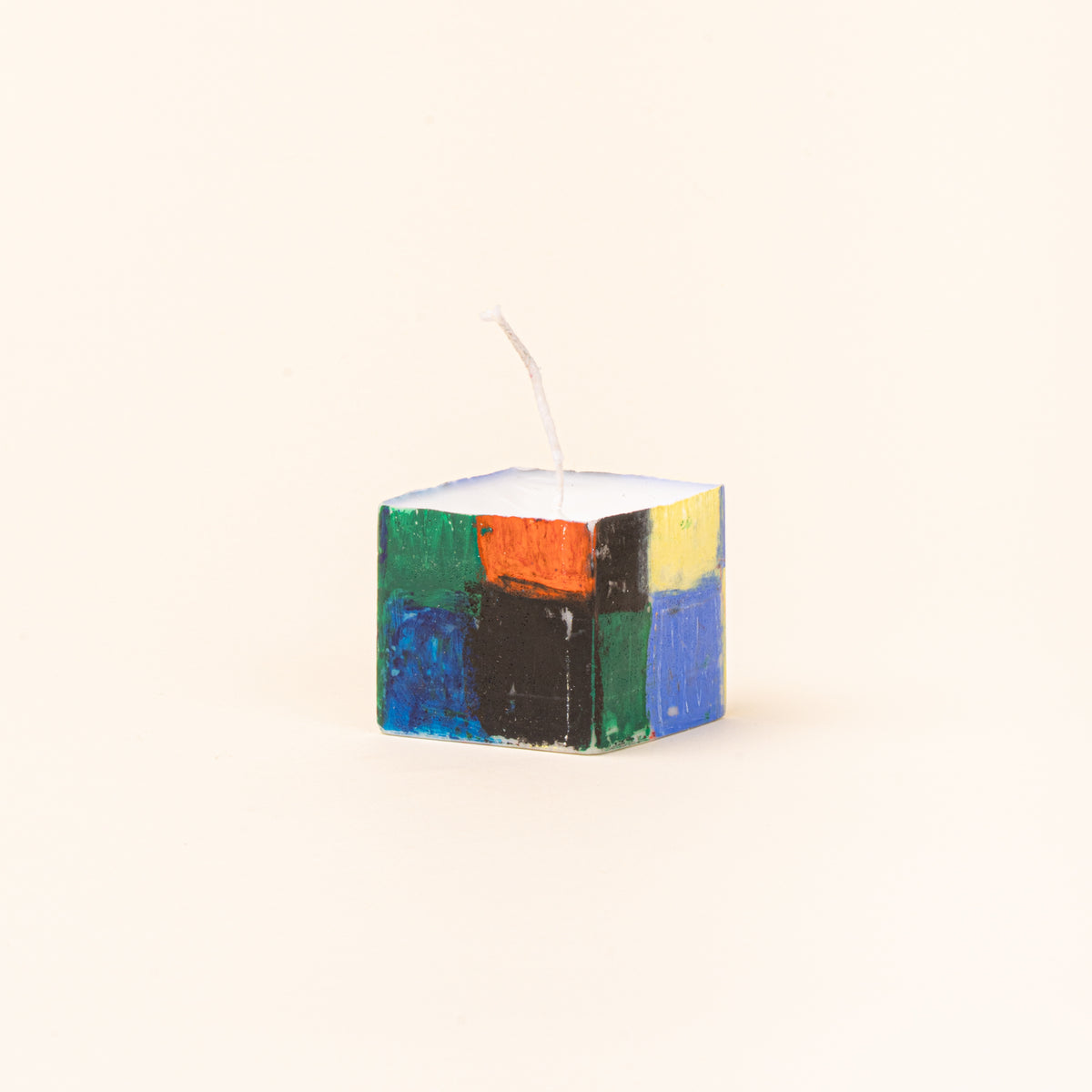 A tall, hand-painted rectangular candle shown beside its original green-and-white checkerboard paper packaging. The candle features blocks of cobalt blue, red, green, yellow, and black, with a white wick at the top.