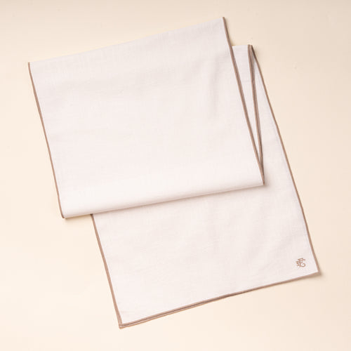 A folded white table runner with taupe-colored piping and embroidered monogram.