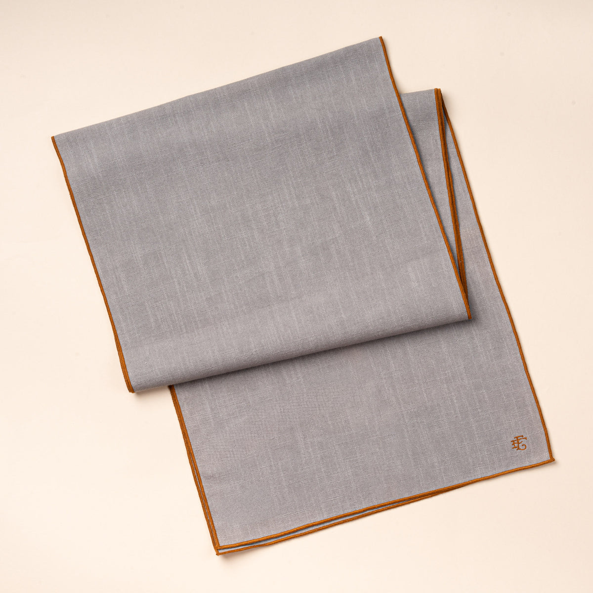 A folded light blue table linen runner with rust-colored piping and embroidered monogram.
