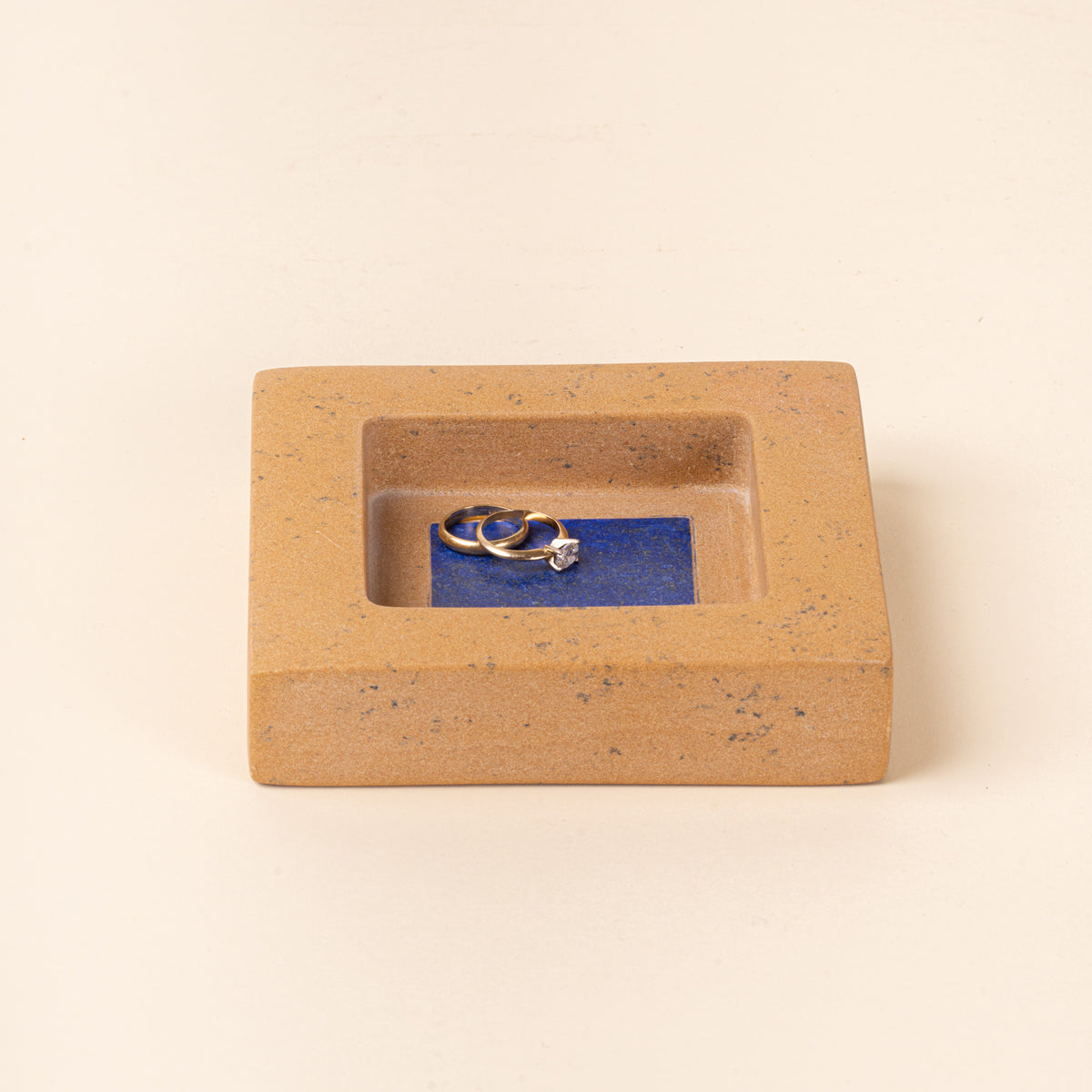 Rectangular tan speckled jewelry dish with a recessed center and blue interior, holding two gold rings.