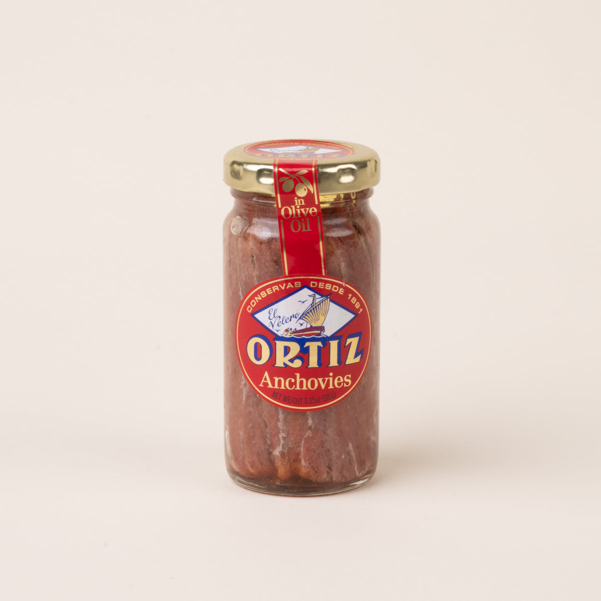 Glass jar of Ortiz anchovies in olive oil with a gold lid and red, blue, and gold label on a cream background.