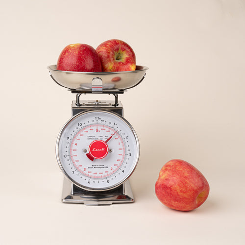 Stainless steel vintage kitchen scale with two red apples in the weighing bowl and one apple beside it.