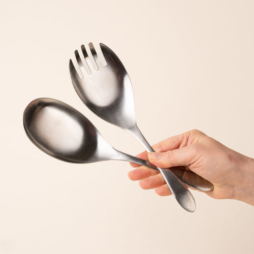A hand holds a stainless-steel salad serving set with an oversized spoon and four-tined serving fork, featuring smooth oval heads and gently tapered handles, photographed on a warm cream background.