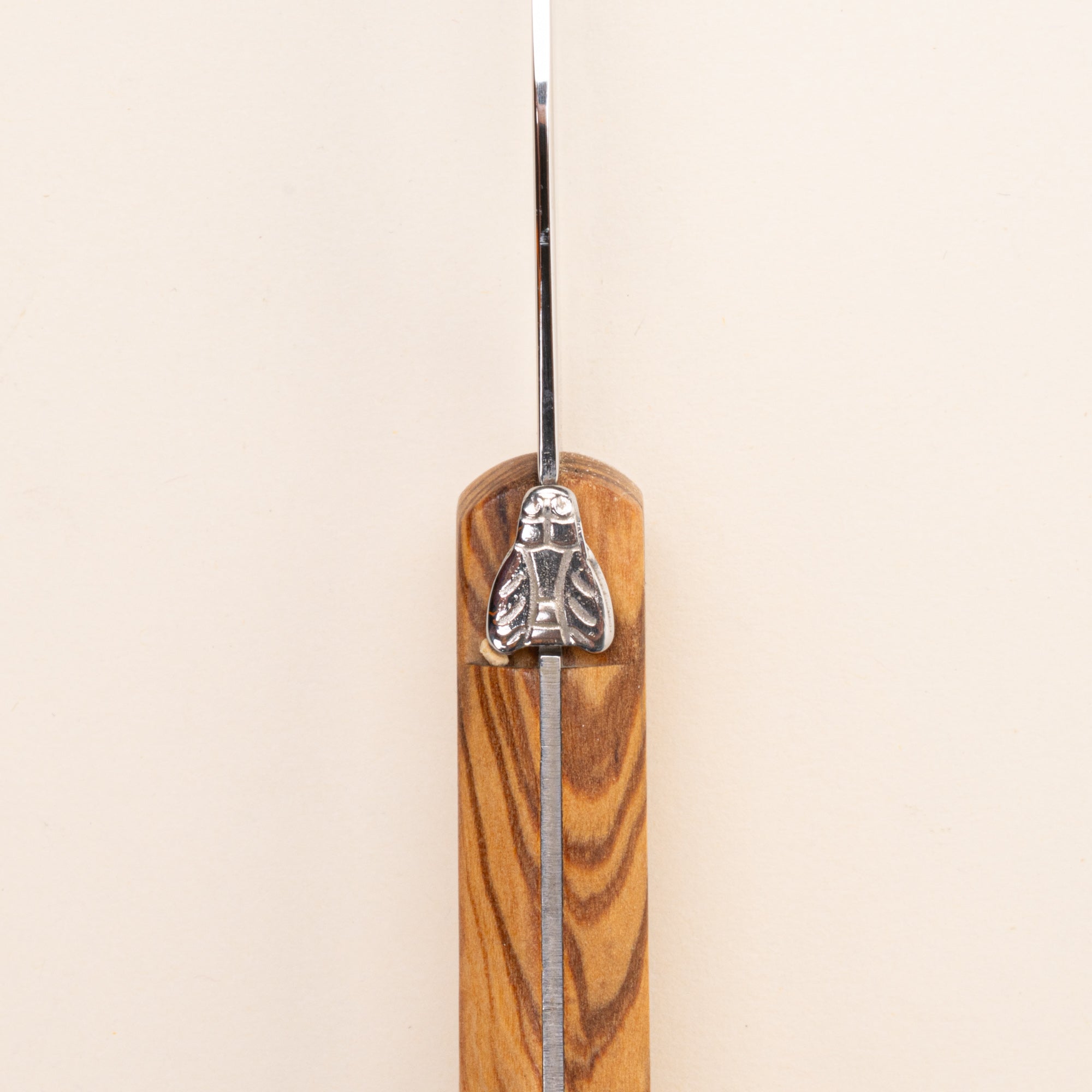 A close-up of a knife handle made of warm-toned olive wood with a polished silver metal blade and a decorative silver bee emblem on the guard.
