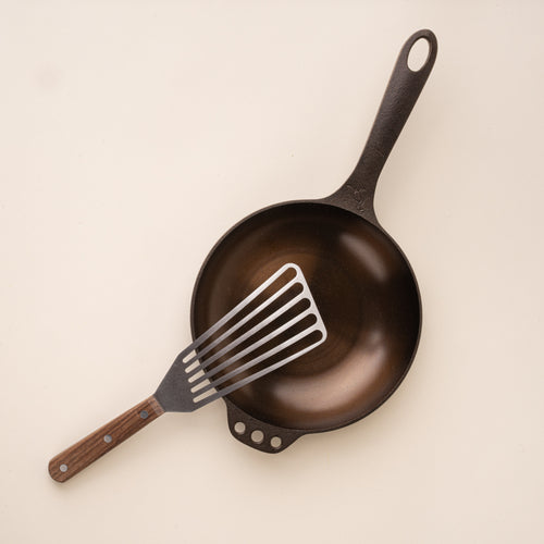 Slotted stainless steel spatula with a wooden handle, resting on a small cast iron pan.