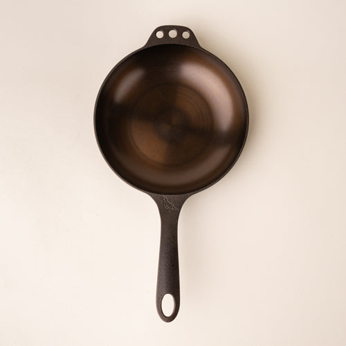 A dark brown small cast iron skillet with a smooth cooking surface and three holes on the opposite side, with a handle.