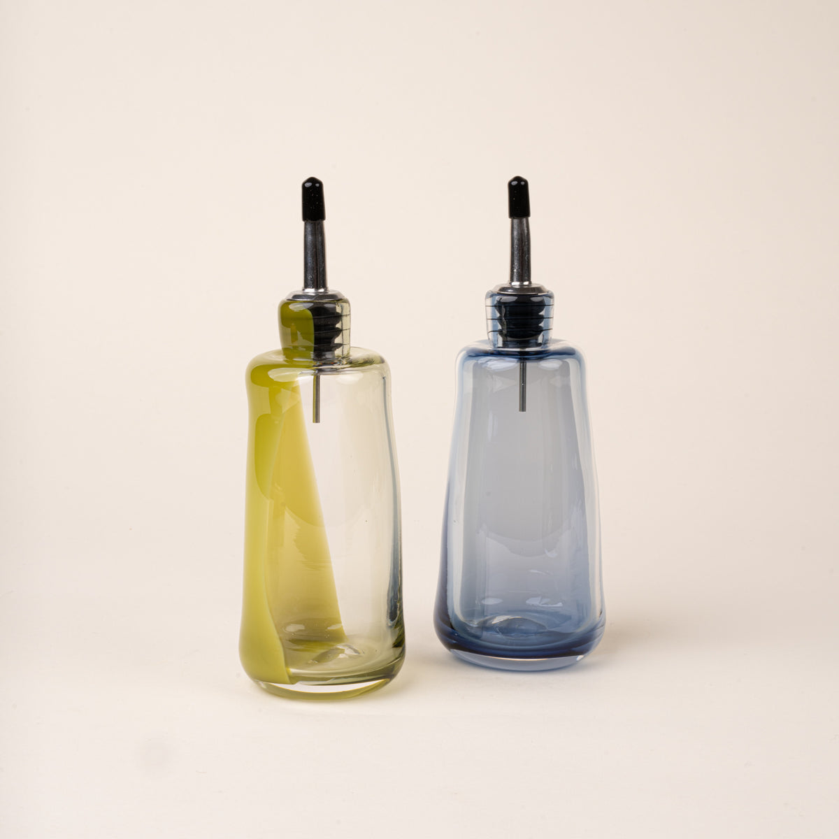 An olive-green and blue-grey glass oil or vinegar dispensers with a polished metal pour spout and black stopper.