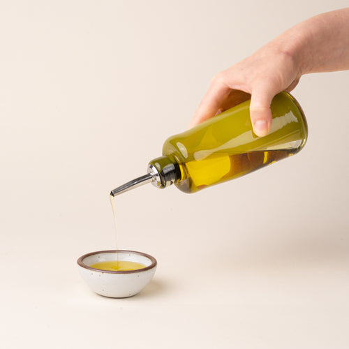 A hand holds an olive-green glass oil or vinegar dispenser with a polished metal pour spout and black stopper, pouring oil into a small ceramic white bowl.