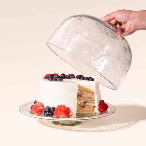 A hand lifts an etched floral glass dome from a berry-topped layer cake on a clear glass cake stand.