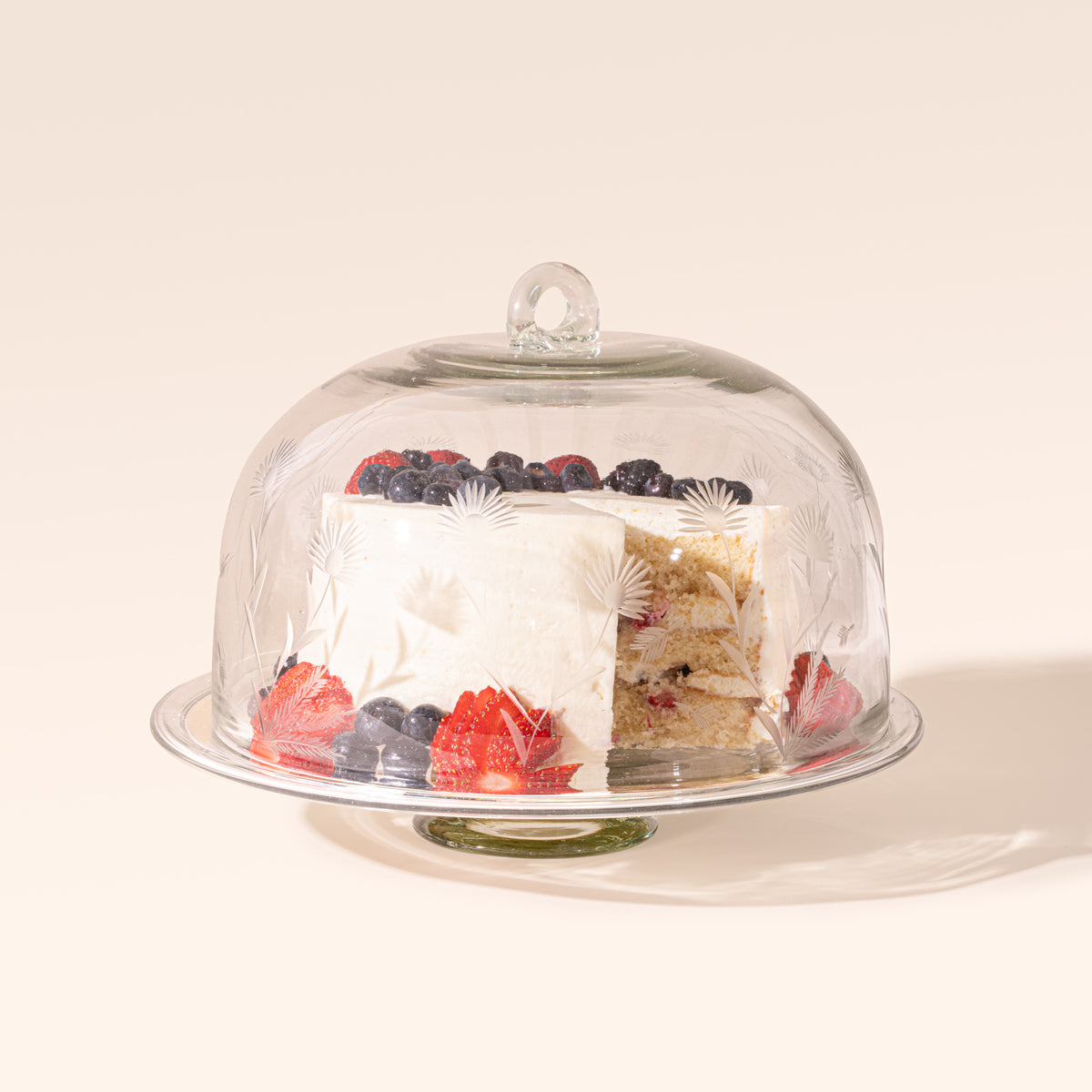 An etched floral glass dome on a clear glass cake stand with a berry-topped layer cake inside.