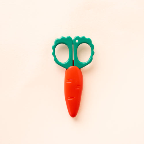 Carrot-shaped scissors with green scalloped handles and an orange protective blade cover, closed against a cream background.