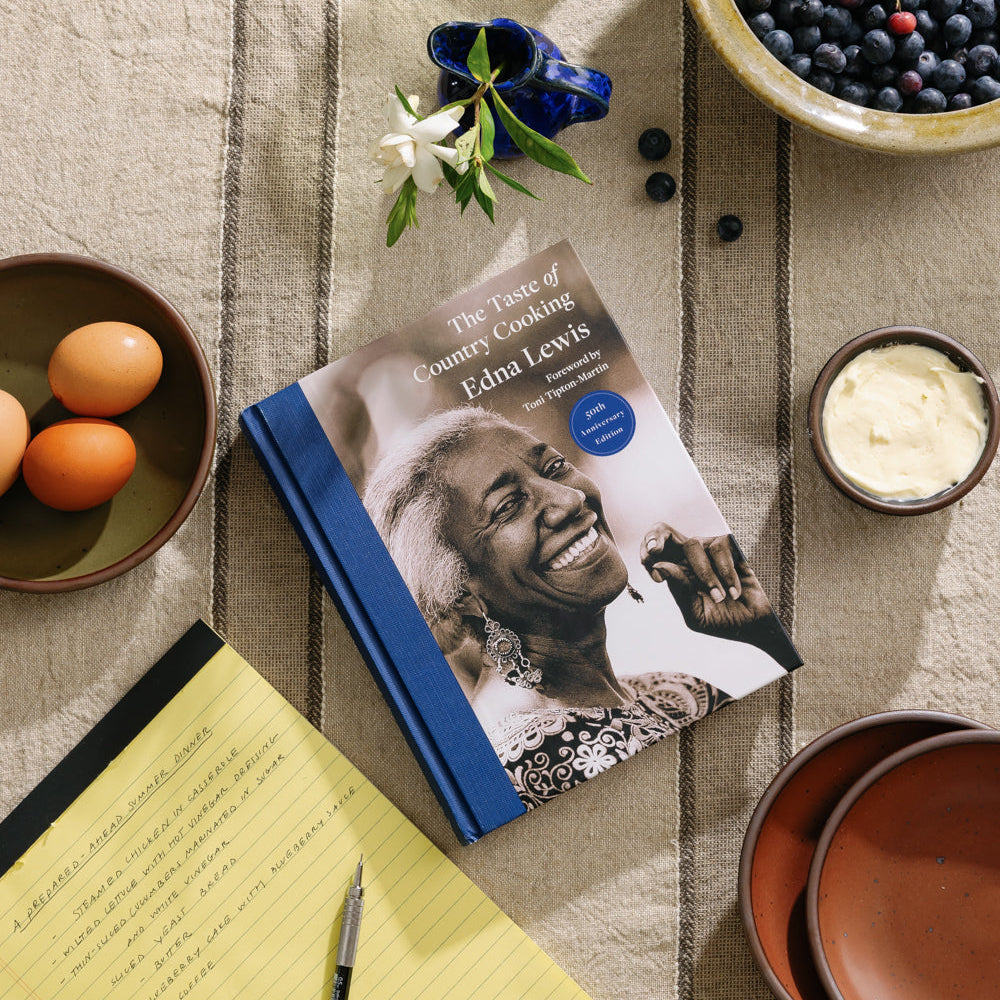 The cookbook, The Taste of Country Cooking, by Edna Lewis, sits on a tablecloth surrounded by a yellow legal pad and pen, ceramic dinnerware with ingredients, and a vase with flowers.