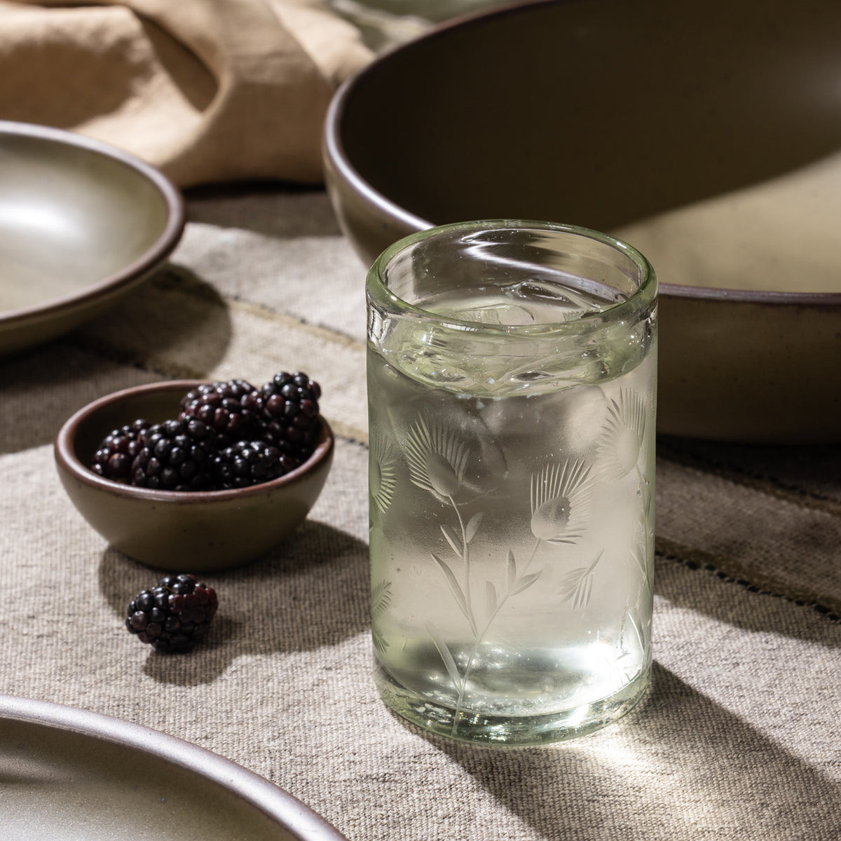 Etched glass tumbler filled with ice and a clear beverage beside a small bowl of blackberries and ceramic dinnerware.