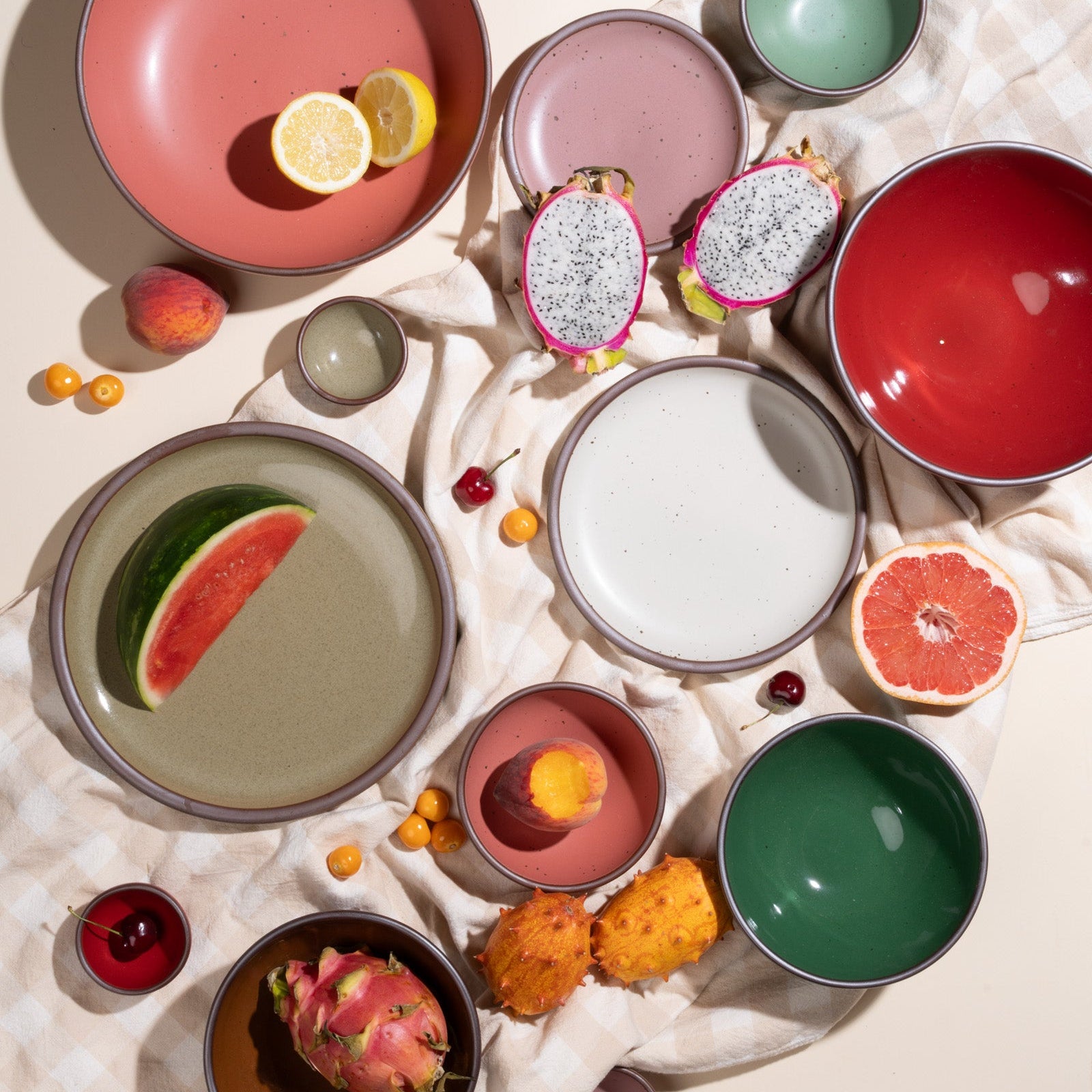 Overhead view of ceramic plates and bowls in a variety of colors arranged with a picnic blanket with fruits.