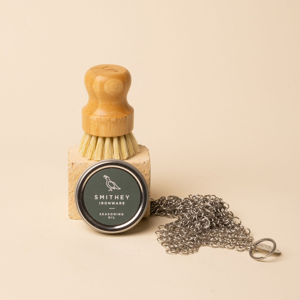 Smithey Ironware seasoning oil tin, wooden cleaning brush, and chainmail scrubber on neutral background.