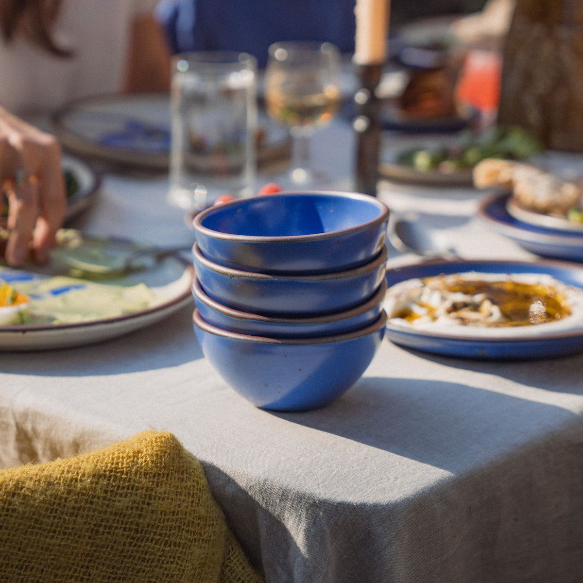 On an outdoor table is a stack of 4 small dessert sized rounded ceramic bowls in a cobalt blue color featuring iron speckles and an unglazed rim, along with other dishes and foods.