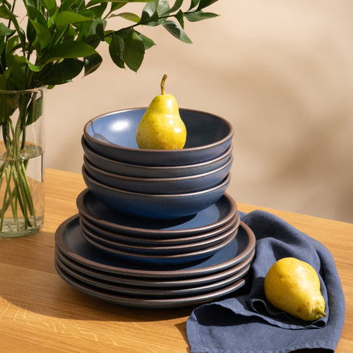4 ceramic dinner plates, 4 side plates, and 4 shallow dinner bowls in a cool medium blue color with iron speckles and unglazed rims are stacked all together on a table with pears.