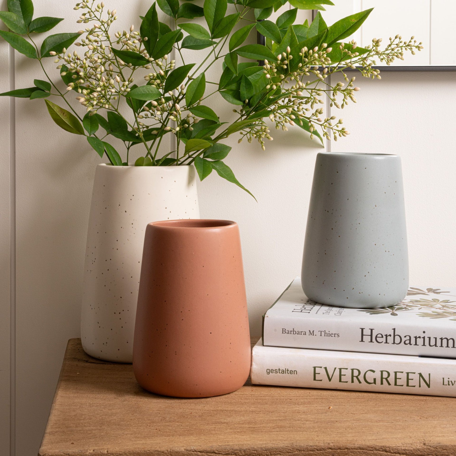 Three speckled vases in ivory, terracotta, and blue arranged beside greenery and stacked books on a wooden surface.