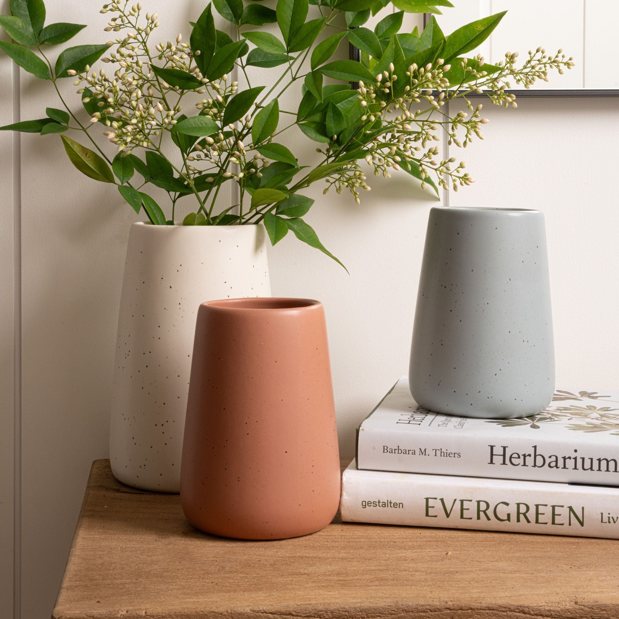 Three speckled vases in ivory, terracotta, and blue arranged beside greenery and stacked books on a wooden surface.