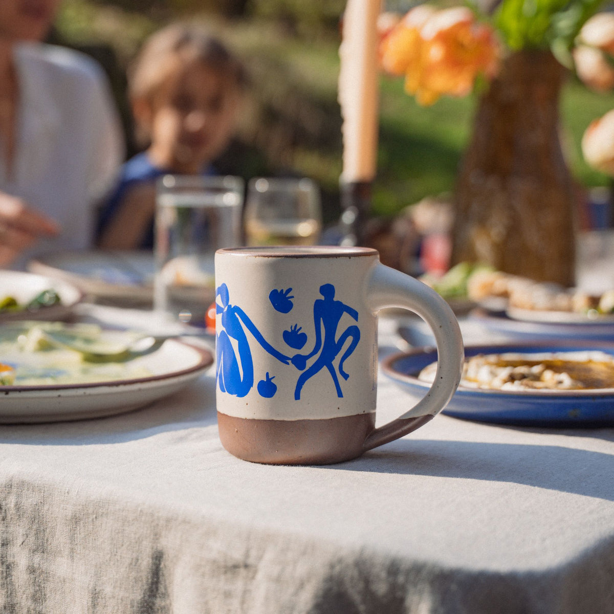 On an outdoor table, sits East Fork’s The Mug in a warm off white color with handle with a blue decal of Henri Matisse’s Femmes et singes, and a unglazed rim and bottom base, sits on a table with gold fabric and oranges.