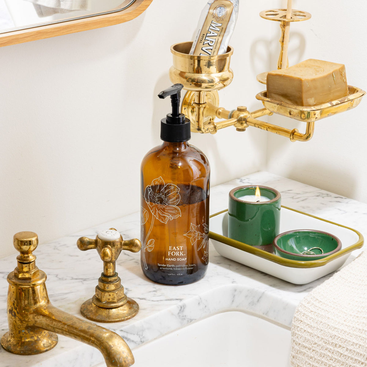 Amber hand-soap bottle on a marble sink beside brass fixtures, a green ceramic candle, and a tray with small green dishes in a bright bathroom.