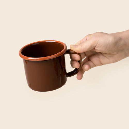 A hand holds out a red-brown enamel mug with an orange rim and handle, shown against a neutral beige background.