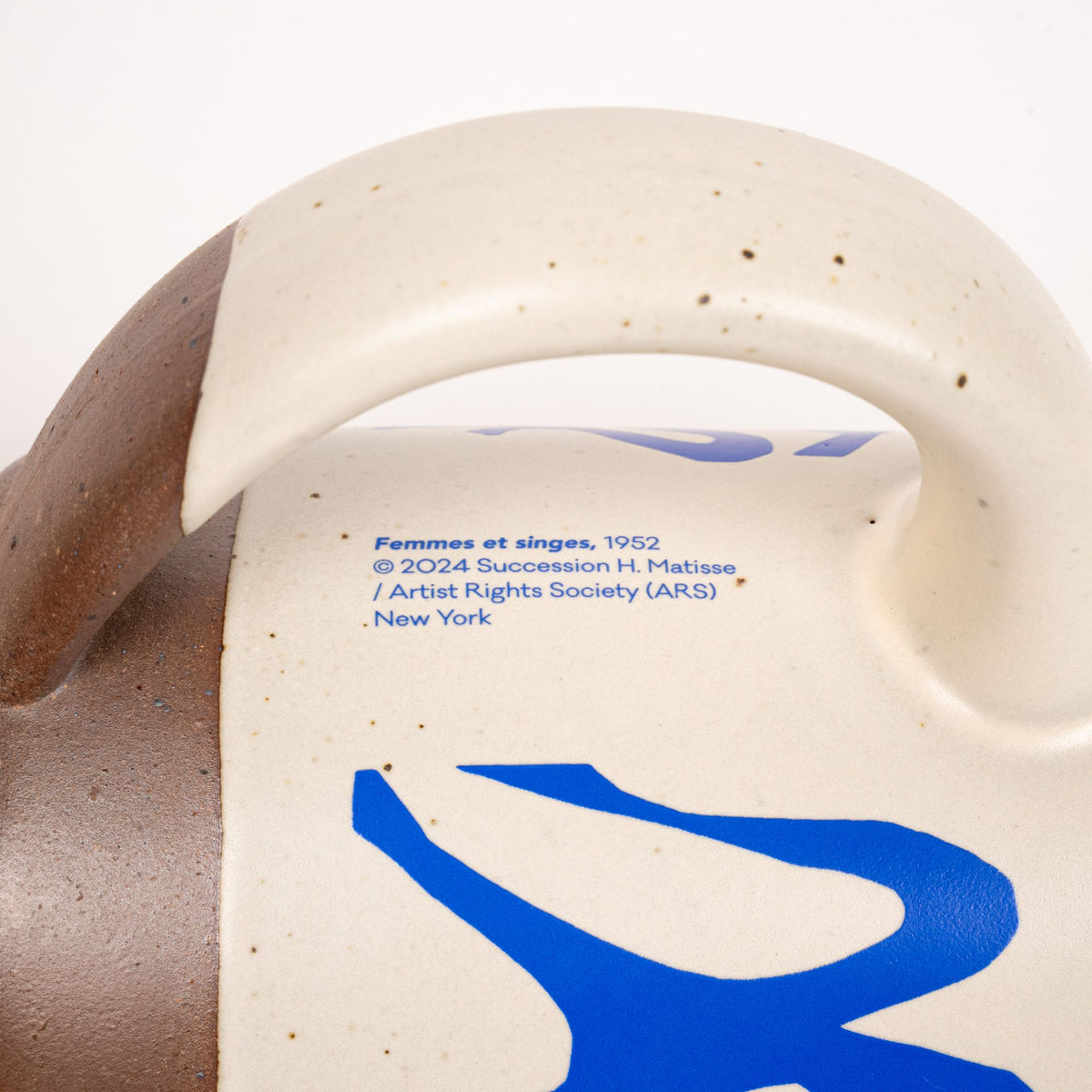 A closeup of the copyright on East Fork’s The Mug in a warm off white color with handle with a blue decal of Henri Matisse’s Femmes et singes, and a unglazed rim and bottom base.