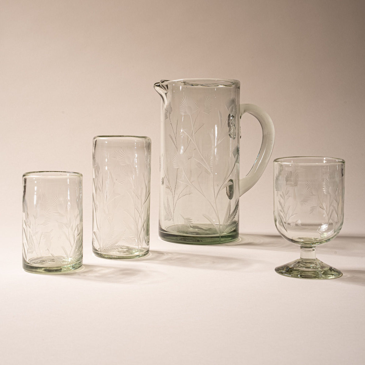 Set of clear etched glassware with floral design, including pitcher, wine glass, highball glass, and short tumbler on neutral background.