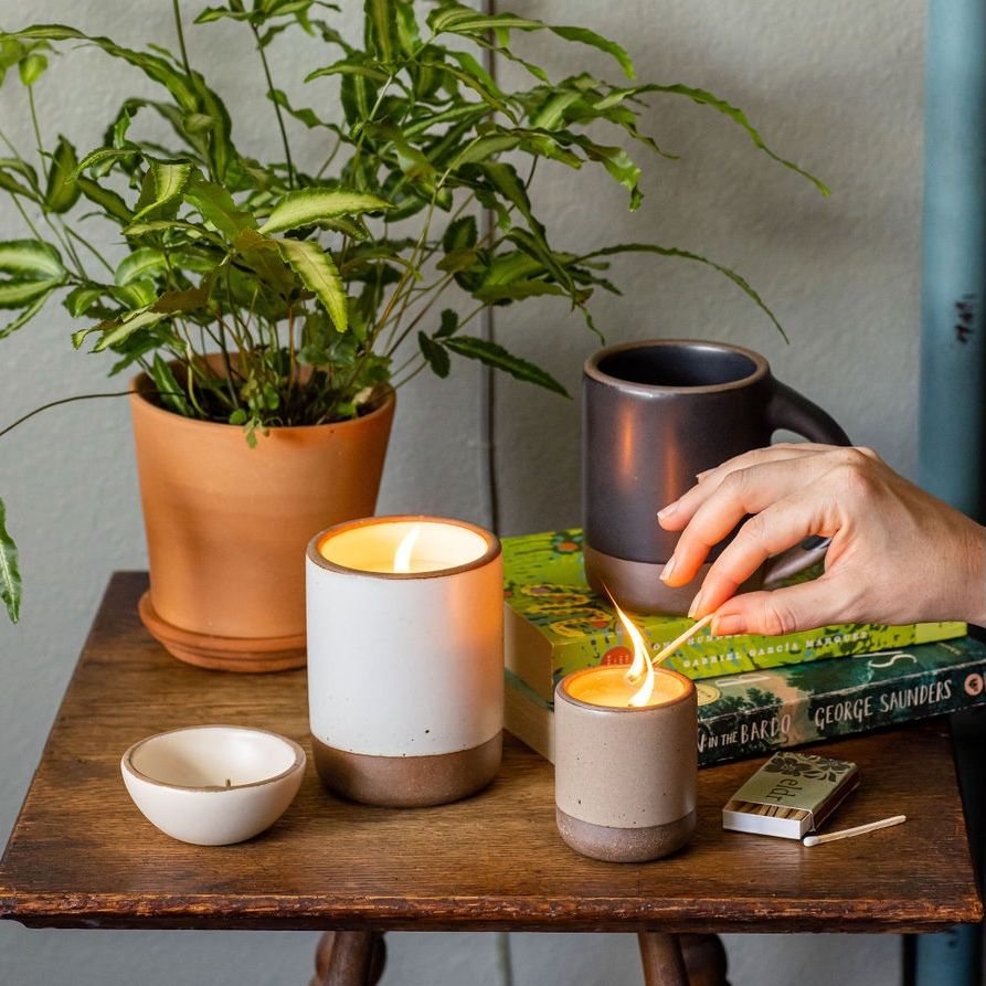 On a nightstand, a hand is lighting a candle in a ceramic vessel. There's a larger candle already lit, a small tiny bowl, a ceramic mug on a stack of books, and a potted plant.