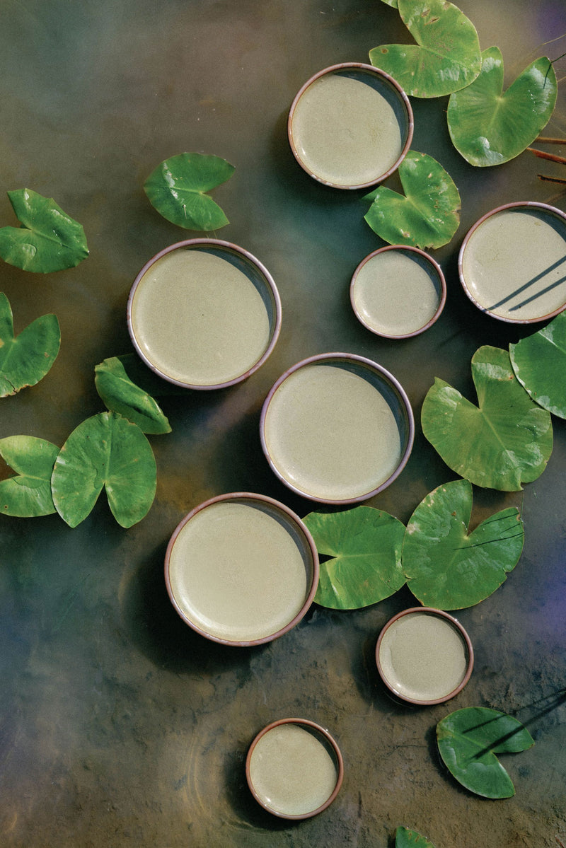 Ceramic bowls with a light green glaze are artfully arranged on a water surface amidst green lily pads, creating a serene, natural composition.