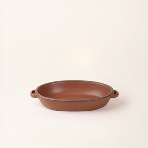 A medium oval, matte cool terracotta ceramic baking dish with two side handles, on a clean, solid off-white background.