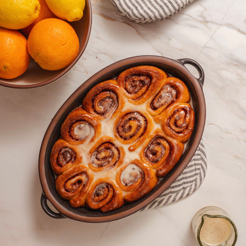 An overhead view of a medium oval, graphite black, ceramic baking dish with handles, filled with golden, glazed cinnamon rolls surrounded by a bowl of oranges and striped pot holders. 