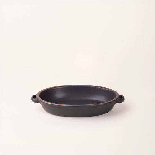 A medium, oval, matte, graphite black ceramic baking dish with two side handles, on a solid off-white background.
