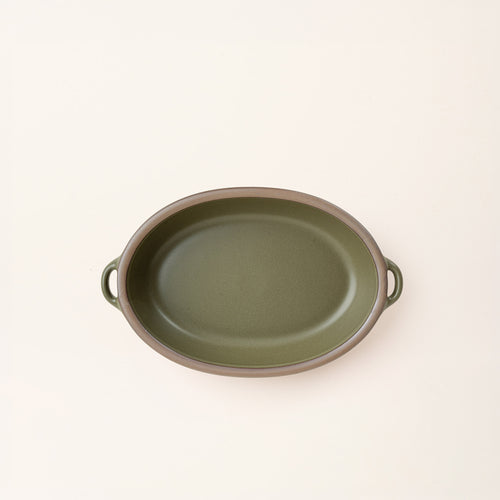 A medium, oval, matte, olive green ceramic baking dish with two side handles, on a solid off-white background, photographed from overhead. 