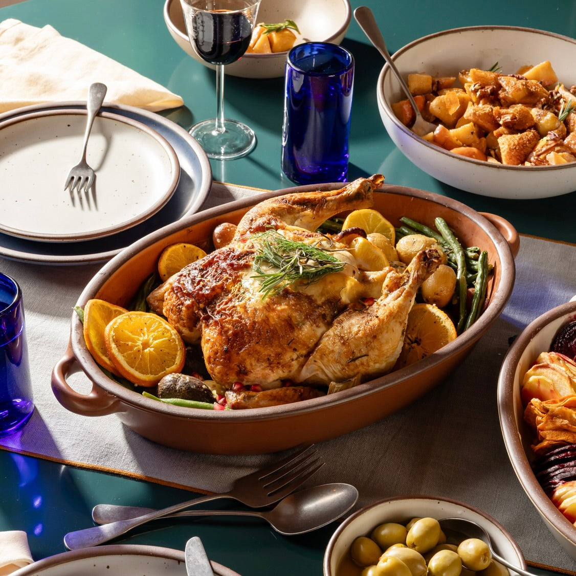 An overhead view of a large oval, terracotta colored, ceramic baking dish with handles, filled with a roast chicken, potatoes and green beans, with orange slice garnish. The disc is surrounded by ceramic bowls, plates dark blue and steel flatware. 